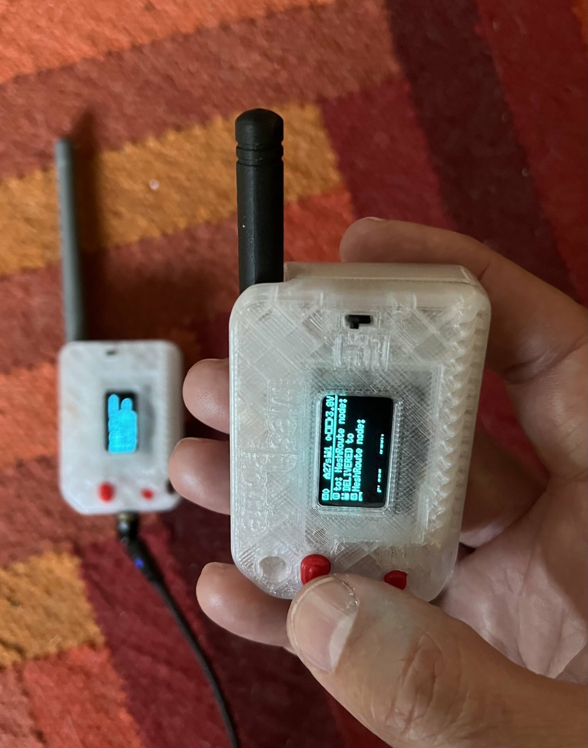 Hand holding a transparent-cased Heltec V3 MeshRoute device with its OLED screen lit; a second radio is visible behind it.
